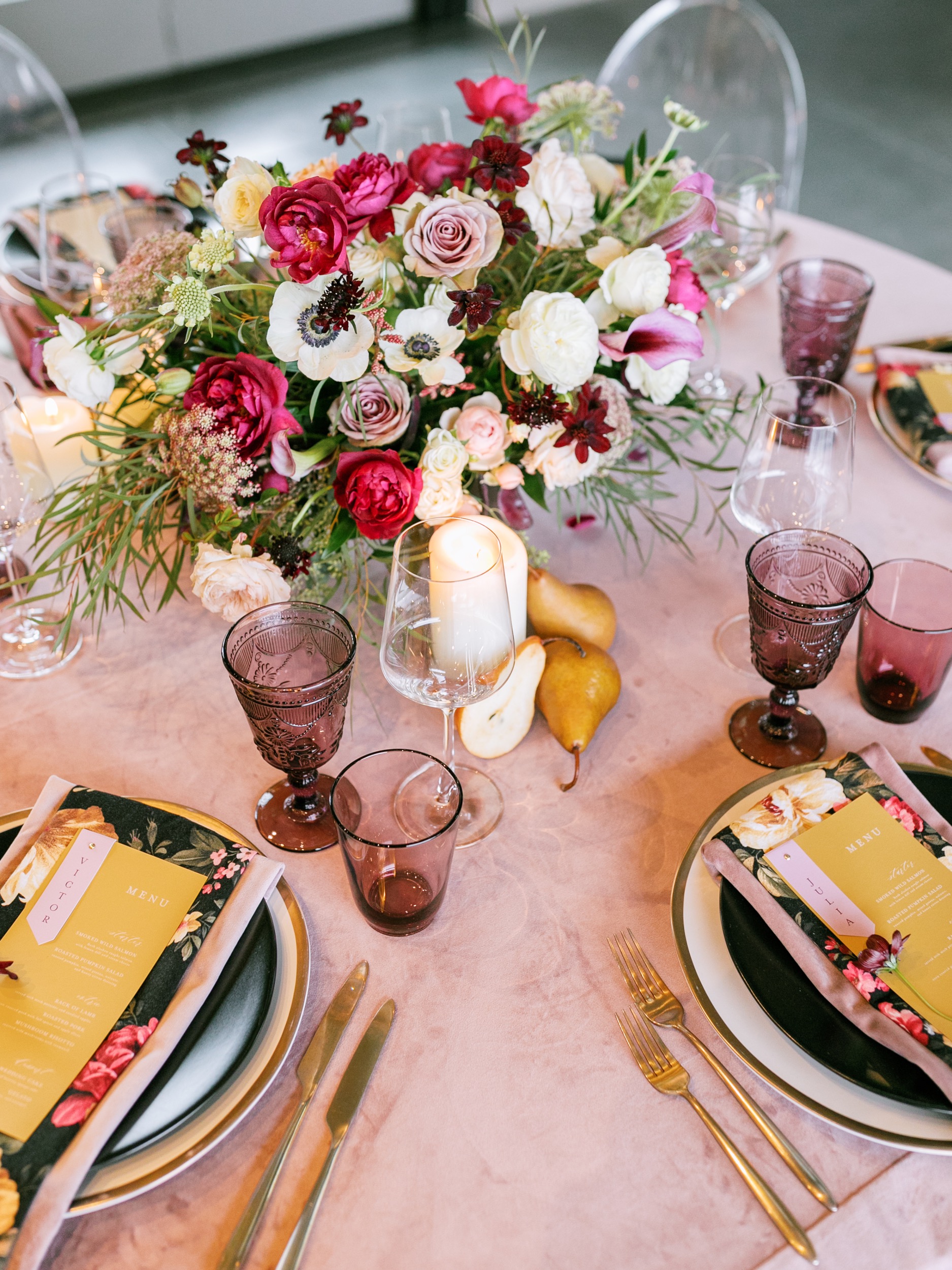Blush and Romantic Wedding Table with White and Gold Plates topped with Modern Black Plates, Sleek Gold Flatware and an Untamed Centerpiece with Cream and Deep Burgundy Flowers Surrounded by Pears and Candles